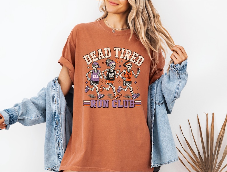 May include: A rust-colored short-sleeve t-shirt featuring a graphic of three skeletons running. The text reads DEAD TIRED RUN CLUB. The skeletons wear athletic gear with text including 13.1, Last Mile, and Best in Pace. The design uses orange, purple, and white.