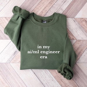 Peut inclure: Un sweat-shirt vert avec le texte "in my ai/ml engineer era" imprimé en blanc.