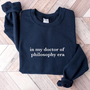 May include: A navy blue sweatshirt with the text "in my doctor of philosophy era" printed in white on the front.