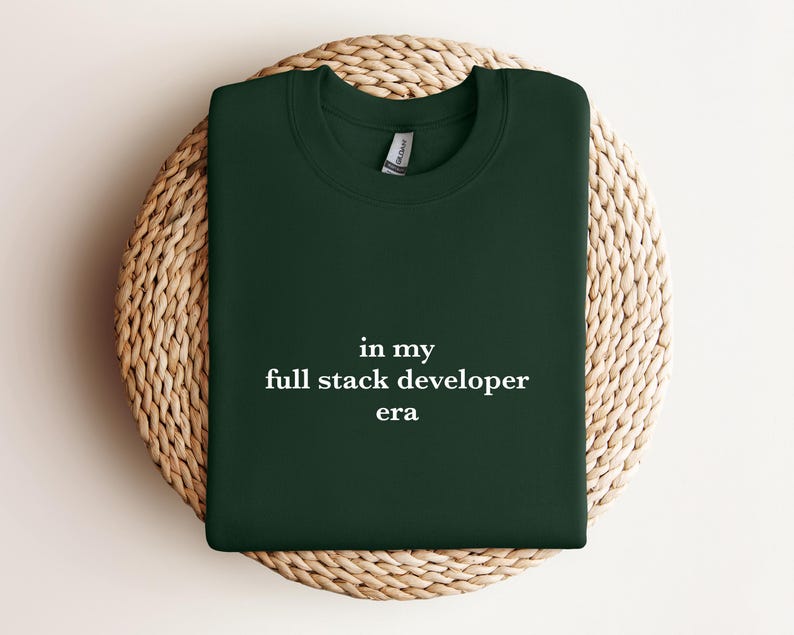 A dark green t-shirt with the text "in my full stack developer era" printed on it, laid out on a woven basket.