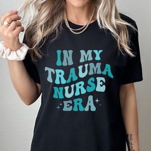 Pu&ograve; includere: Maglietta nera con il testo "IN MY TRAUMA NURSE ERA" in un font degradato blu e turchese con stelle su entrambi i lati del testo.