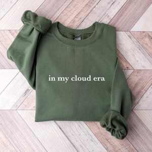 May include: An olive green crewneck sweatshirt featuring the white text "in my cloud era" printed across the center of the chest. The garment has long sleeves with ribbed cuffs and a ribbed neckline, constructed from a solid-colored fabric.