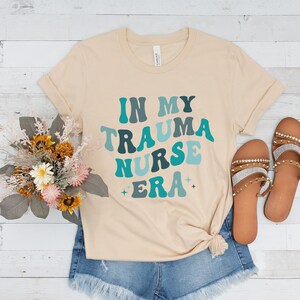 Pu&ograve; includere: Una maglietta beige con il testo "In My Trauma Nurse Era" in un font retr&ograve; con colori blu e verdi.