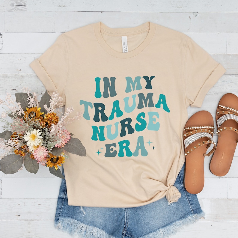 Pu&ograve; includere: Una maglietta beige con il testo "In My Trauma Nurse Era" in un font retr&ograve; con colori blu e verdi.