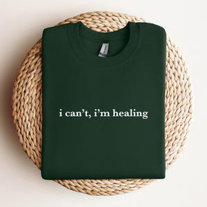 A dark green t-shirt with the text "I can't, I'm healing" printed on it, laid out on a woven basket.