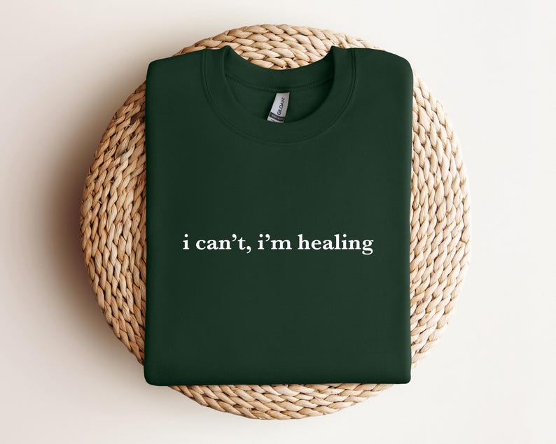 A dark green t-shirt with the text "I can't, I'm healing" printed on it, laid out on a woven basket.