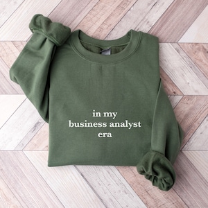May include: A green sweatshirt with the text "in my business analyst era" printed in white on the front.