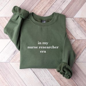 May include: A dark green sweatshirt with the text "in my nurse researcher era" printed in white on the front.