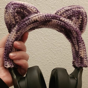 May include: Purple and white crocheted cat ear headband over black headphones.