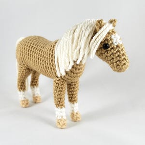 Puede incluir: Un caballo de crochet marrón con una crin blanca y marcas blancas en sus patas y cara. El caballo tiene ojos negros y una nariz marrón.