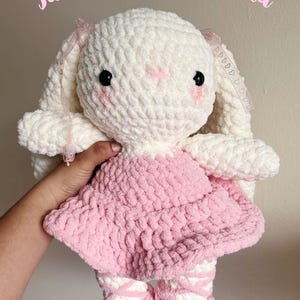 May include: A crocheted white bunny ballerina doll wearing a pink dress and white shoes. The doll is holding a pink ribbon in its hand. The doll is a PDF pattern.