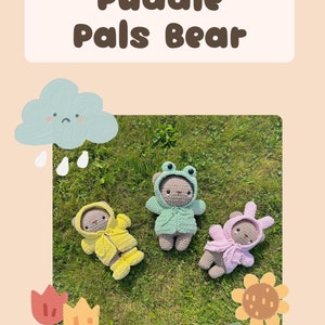 May include: Three crocheted teddy bears wearing different outfits: a yellow raincoat, a green frog costume, and a pink bunny costume. The bears are lying on green grass with a cloud and raindrops above them.