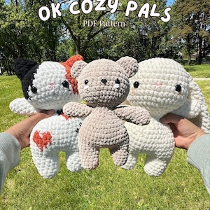 Puede incluir: Un grupo de animales de peluche de ganchillo hechos a mano, incluyendo un gato, un oso y un conejo. El gato es blanco con detalles negros y naranjas, el oso es marrón y el conejo es blanco. El texto "OK COZY PALS PDF Pattern" está en la parte superior.