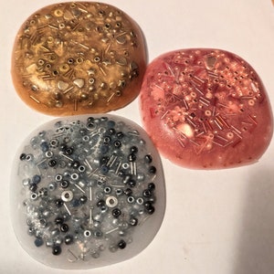 Può includere: Tre sottobicchieri rotondi in resina con sfondi di colori diversi e varie piccole perline e paillettes incastonate nella resina. I sottobicchieri sono in tonalità dorate, rosa e trasparenti con perline nere.