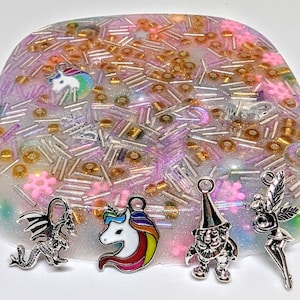 Può includere: Un vassoio in resina trasparente pieno di perline colorate e glitter, con vari ciondoli attaccati al vassoio, tra cui un unicorno, un drago, un gnomo e una fata.