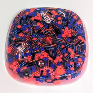 May include: A round, pink resin coaster with a spiderweb design and red, blue, and black sprinkles. A small, black and red Spider-Man logo is visible in the center.