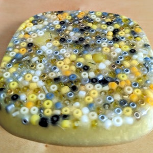 Può includere: Un sottobicchiere rotondo in resina trasparente con un mosaico di piccole perline colorate. Le perline sono principalmente gialle, bianche e nere, con alcune perline blu e argentate mescolate.
