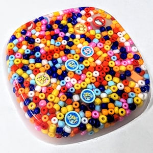 May include: A round, clear resin coaster with a colorful mix of red, orange, yellow, blue, and white beads. The coaster features various cartoon characters, including a Pikachu, a Squirtle, and a Bulbasaur.