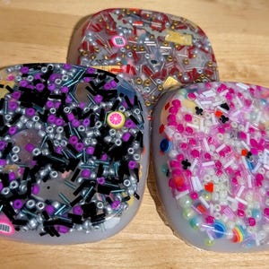 May include: Three resin coasters with embedded decorative elements. One coaster features black and silver beads with purple accents and fruit slices. Another has red, gold, and silver beads. The third has pink and white beads.