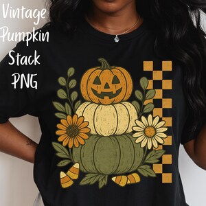 Puede incluir: Camiseta negra con un diseño de calabazas apiladas. La calabaza superior tiene una cara tallada, la del medio es amarilla y la inferior es verde. El diseño incluye girasoles, hojas y el texto "Vintage Pumpkin Stack PNG".