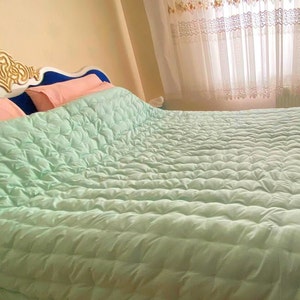 May include: A light green quilted bedspread with a textured surface. The bedspread is on a bed with a blue headboard and pink pillows.
