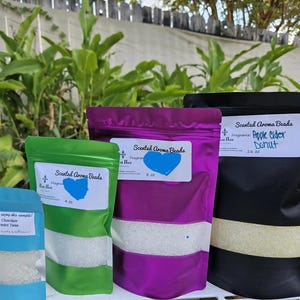 May include: Four colourful pouches of scented aroma beads. The pouches are blue, green, purple, and black, each with a white label. The labels display the scent and weight, including "Apple Cider Donut" and "Chocolate Peppermint Twist."