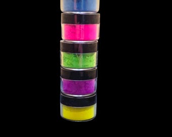 May include: A stack of five small jars filled with colorful powder. The jars are clear with black lids. The powders are blue, pink, green, purple, and yellow.