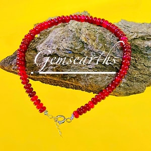 May include: A red beaded necklace with a silver clasp. The necklace is displayed on a piece of wood against a yellow background. The text "Gemsearths" is written in white cursive on the wood.