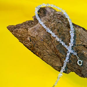 Peut inclure: Un collier délicat composé de petites perles claires de forme irrégulière. Le collier est présenté sur un morceau de bois brun texturé sur un fond jaune vif. Il possède un fermoir argenté.