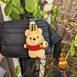 Puede incluir: Un amuleto de ganchillo hecho a mano de Winnie the Pooh con un tarro de miel y detalles de abeja, colocado en una bolsa negra. El amuleto es amarillo, rojo y marrón. Una abeja de dibujos animados está en el fondo.