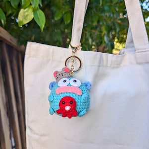 May include: A handmade blue crochet keychain featuring a whimsical monster design with large white eyes, a pink mouth, and a red octopus. A small gray cat is perched on top. The keychain is attached to a gold-colored ring.