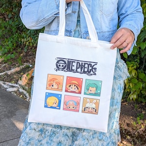 Op de afbeelding: Witte canvas tote bag met de tekst "ONE PIECE" en een logo van een schedel en gekruiste botten. Daaronder zes kleurrijke vierkanten met anime-personage-illustraties. De tas heeft twee handvatten en wordt vastgehouden.