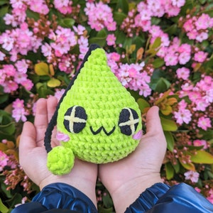 Puede incluir: Un juguete de peluche verde tejido a crochet con forma de l&aacute;grima, con ojos negros y una boca rosa. El juguete est&aacute; siendo sostenido en las manos de una persona.