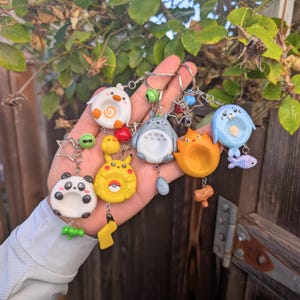Può includere: Una collezione di cinque portachiavi a tema con un panda bianco, un Pikachu giallo, un Totoro grigio, una creatura di fuoco arancione e una foca blu. Ogni portachiavi include un anello di metallo, un campanellino e un accessorio unico.
