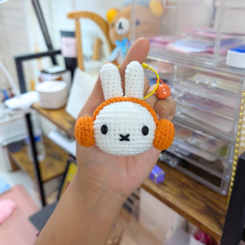 May include: A white crocheted bunny with orange earmuffs. The bunny has black eyes and a black nose. The bunny is being held by a hand.