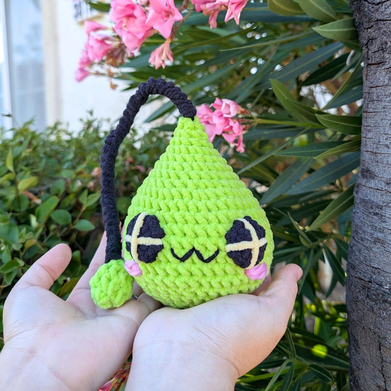 Puede incluir: Un juguete de peluche verde de crochet con forma de l&aacute;grima y una cola de hilo negro. El juguete tiene una cara sonriente con ojos negros y mejillas rosadas.
