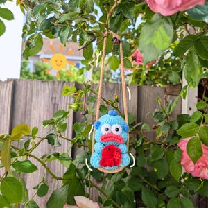 May include: A handmade crochet toy in the shape of a blue monster with a red heart-shaped belly, hanging from a tree branch. The toy has large white eyes and is suspended by tan rope. The background includes green foliage and pink roses.