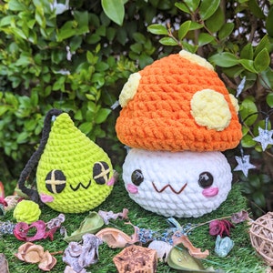Puede incluir: Dos juguetes de peluche de crochet con forma de hongo. El hongo m&aacute;s grande es naranja con manchas amarillas y una cara blanca. El hongo m&aacute;s peque&ntilde;o es verde con una cara blanca. Ambos hongos tienen ojos negros y una boca sonriente.