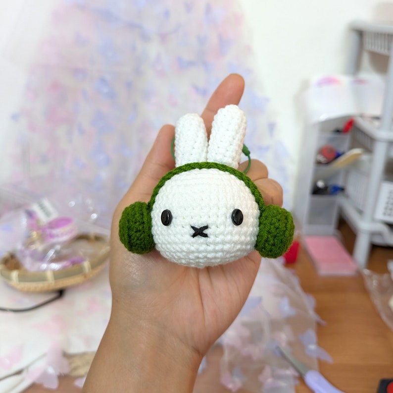 May include: A white crocheted bunny with green crocheted earmuffs. The bunny has black eyes and a black nose.