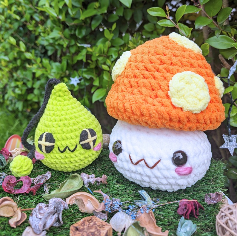 Puede incluir: Dos peluches de crochet, uno con forma de hongo con una cara blanca y detalles de crochet naranja y amarillo, y el otro con forma de l&aacute;grima verde con una cara blanca y negra.