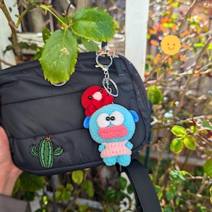 May include: Black quilted crossbody bag with a cactus graphic. A handmade crochet keychain featuring a blue creature with a red hat and a pink sweater is attached to the bag with a silver keyring. The background is a sunny outdoor scene.