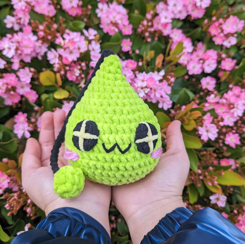 Puede incluir: Un juguete de peluche verde tejido a crochet con forma de l&aacute;grima, con ojos negros y una boca rosa. El juguete est&aacute; siendo sostenido en las manos de una persona.