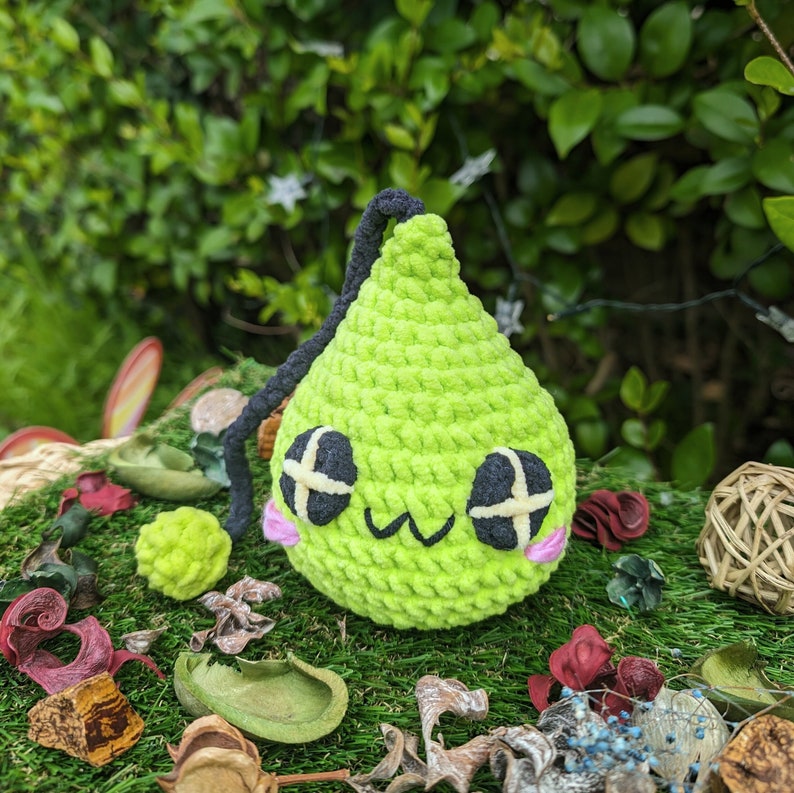 Puede incluir: Un juguete de peluche verde tejido a crochet con forma de pera, ojos negros y mejillas rosadas. El juguete est&aacute; sentado en una cama de musgo verde y otros materiales naturales.