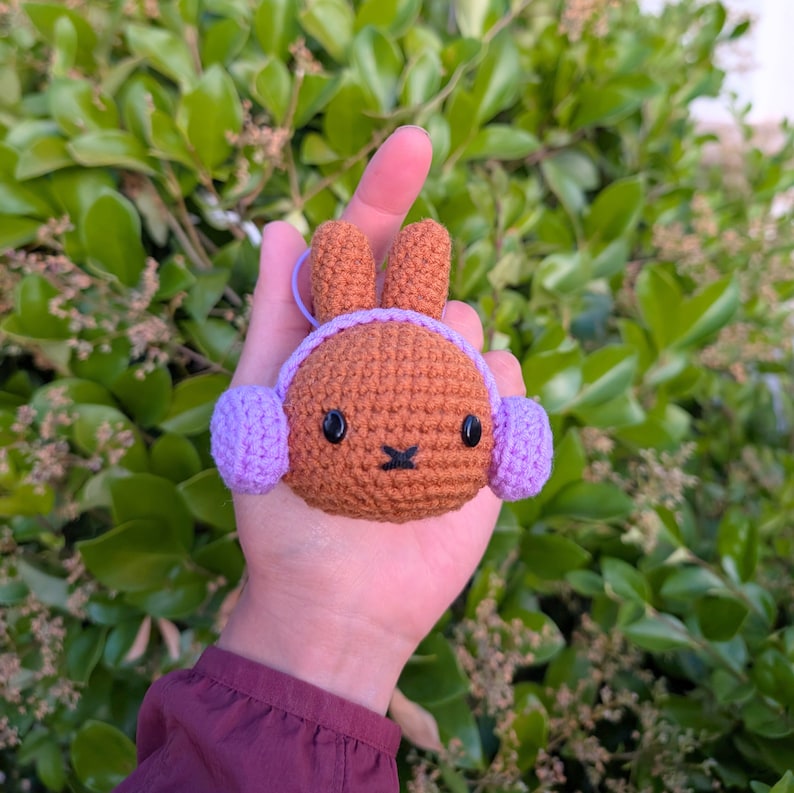 May include: A brown crocheted bunny with purple crocheted earmuffs. The bunny has black eyes and a brown nose.