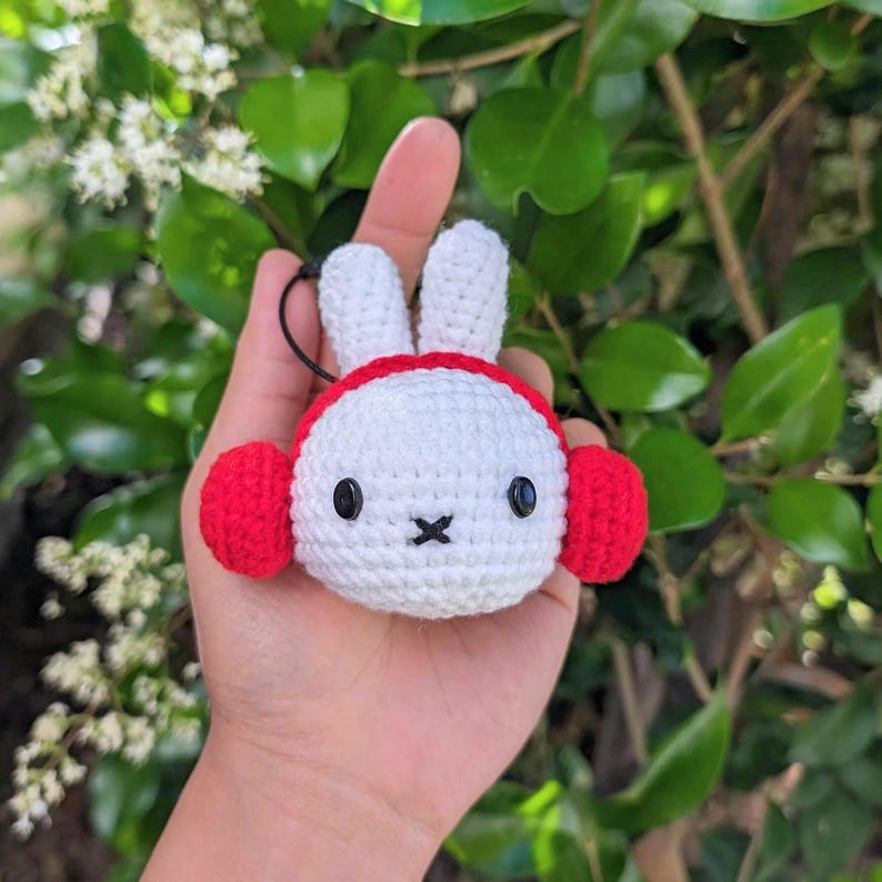May include: White crocheted bunny with red earmuffs. The bunny has black eyes and a black nose.