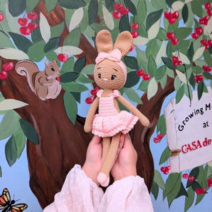 May include: A handmade crochet bunny doll in a pink dress and headband. The doll has long ears and is being held in front of a painted mural of a tree with cherries and a squirrel.