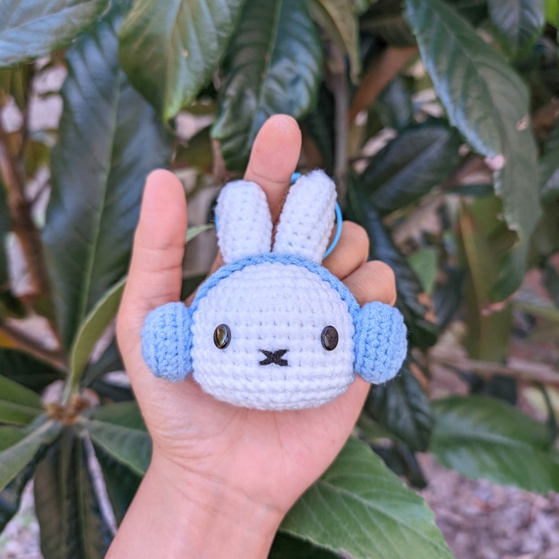 May include: A white crocheted bunny with blue earmuffs. The bunny has black eyes and a small black nose. The bunny is being held by a hand.