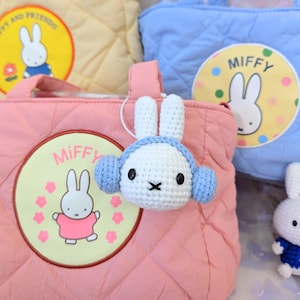 May include: A pink quilted tote bag with a white crocheted bunny wearing blue earmuffs. The bag has a yellow embroidered patch with the word "Miffy" and a cartoon bunny in a pink dress.