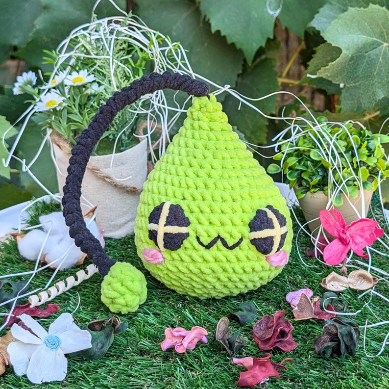 Puede incluir: Un juguete de peluche verde de crochet con forma de l&aacute;grima, ojos negros y una boca sonriente. El juguete tiene una cola de hilo negro.
