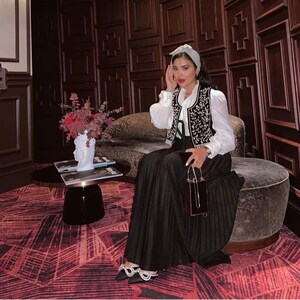 Può includere: Una donna con una camicetta bianca, un gilet nero con ricami floreali e una lunga gonna nera a pieghe. È seduta su un pouf rotondo imbottito. Accessori: fascia per capelli, piccola borsa nera e scarpe decorate.
